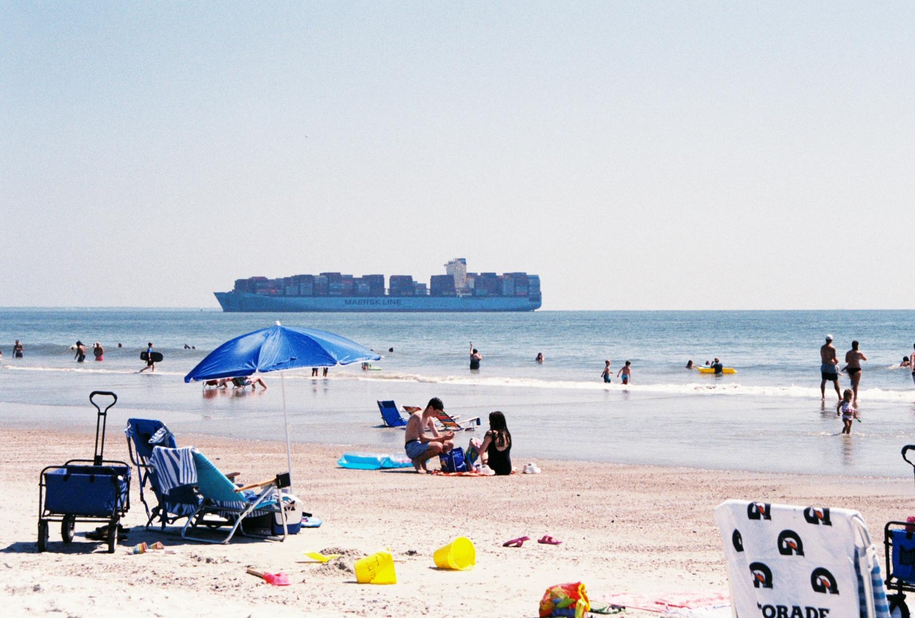 beachgoers on the sand and in the water with a large freight ship in the background