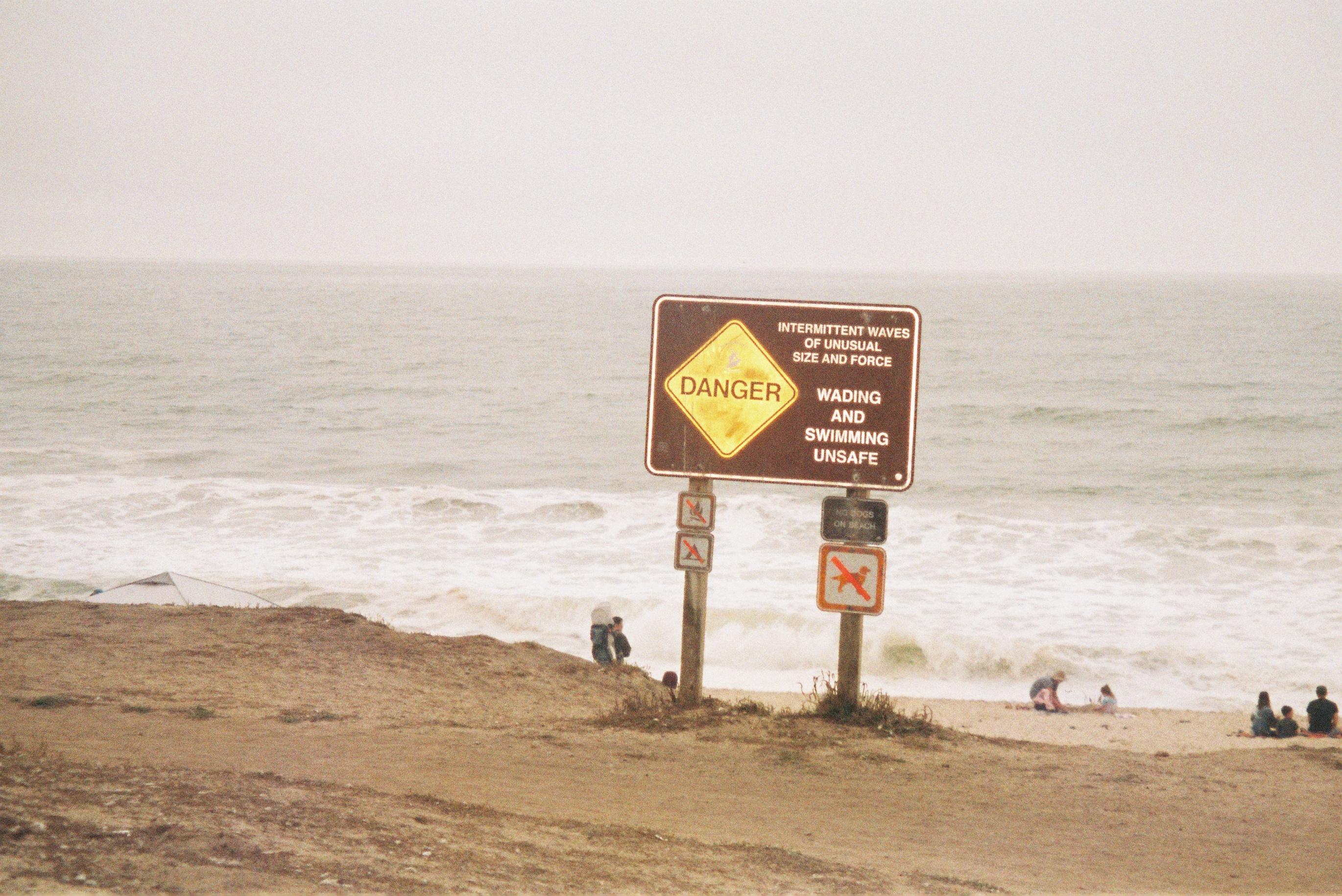 a sign that reads 'DANGER, intermitten waves of unusual size and force, wading and swimming unsafe' on a sandy beach with the ocean in the back
